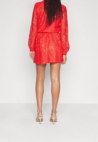 Femme avec de longs cheveux tressés portant une robe courte en dentelle rouge et des sandales à talons beiges, se tenant devant un fond clair et uni.