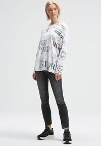 Frau mit kurzen blonden Haaren, die ein weißes Langarmshirt mit schwarzen Textmustern, schwarze Skinny-Jeans und schwarze Sneakers trägt, steht.