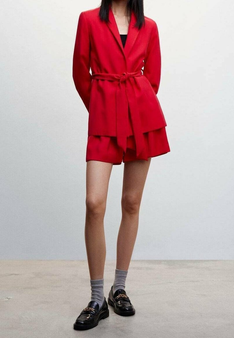 Blazer rojo con shorts a juego, presenta una cintura con cinturón y un diseño de capas. Se lleva con mocasines negros y calcetines grises. Acabado de tela suave.