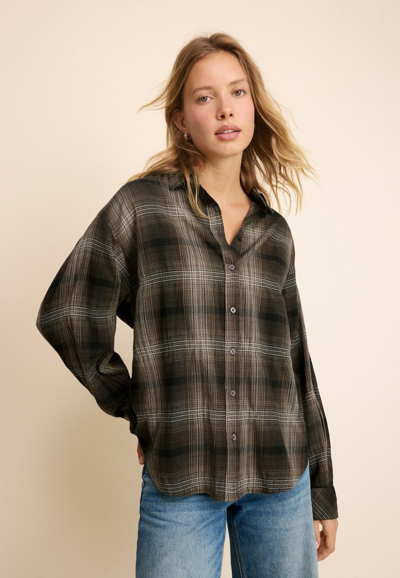 Jeune femme aux cheveux blonds détachés portant une chemise à carreaux marron et noir à boutons et un jean bleu sur un fond beige.