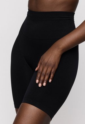 Close-up van een persoon die zwarte biker shorts met hoge taille draagt, met één hand rustend op hun dij tegen een eenvoudige achtergrond.