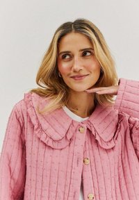 Růžová bavlněná bunda s texturovanou látkou, volánkovým límcem a třemi velkými knoflíky. Má vertikální pruhy a malé vyvýšené tečky po celé ploše.