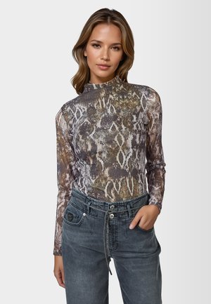 Woman wearing a long-sleeve, high-neck snake print top tucked into high-waisted, faded black jeans with a drawstring waist.