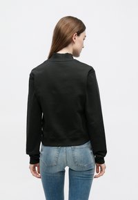 Μαύρο φούτερ με ψηλό γιακά και ριμπ μανσέτες, με εφαρμοστή γραμμή και απαλή υφή. Φορώντας το με μπλε εφαρμοστά τζιν, με έμφαση στην πίσω όψη.