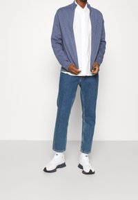 Cardigan blu con zip sopra una camicia bianca a maniche lunghe, abbinato a jeans azzurri a gamba dritta e scarpe sportive bianche con dettagli blu navy.