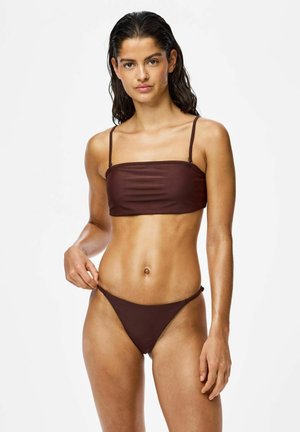 Femme aux cheveux mouillés portant un bikini deux pièces marron foncé, ajustant la bretelle du bas du bikini, debout devant un fond blanc uni.