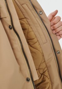 Beżowa kurtka puchowa z pikowanym wnętrzem, z zapięciem na zamek z przodu oraz guzikami. Gładki zewnętrzny materiał z delikatnym połyskiem.