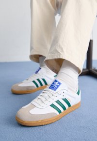 Weiße Sneaker mit grünen Streifen und einem blauen Etikett. Wildleder-Zehenkappe und Kautschuksohle. Getragen mit beigen Hosen und weißen Socken.