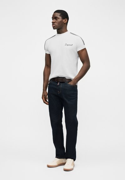 Άνδρας που φοράει λευκό t-shirt Dsquared2, σκούρο τζιν, καφέ ζώνη και λευκά αθλητικά παπούτσια, στέκεται με το ένα χέρι στην τσέπη, κοιτάζοντας αριστερά.