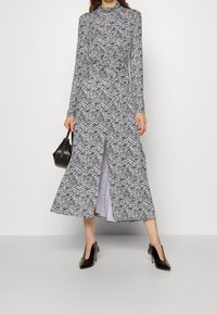 Frau in einem langärmligen Midi-Kleid mit Zebra-Muster und vorderem Schlitz, die eine kleine schwarze Handtasche hält und schwarze Absatzschuhe trägt.