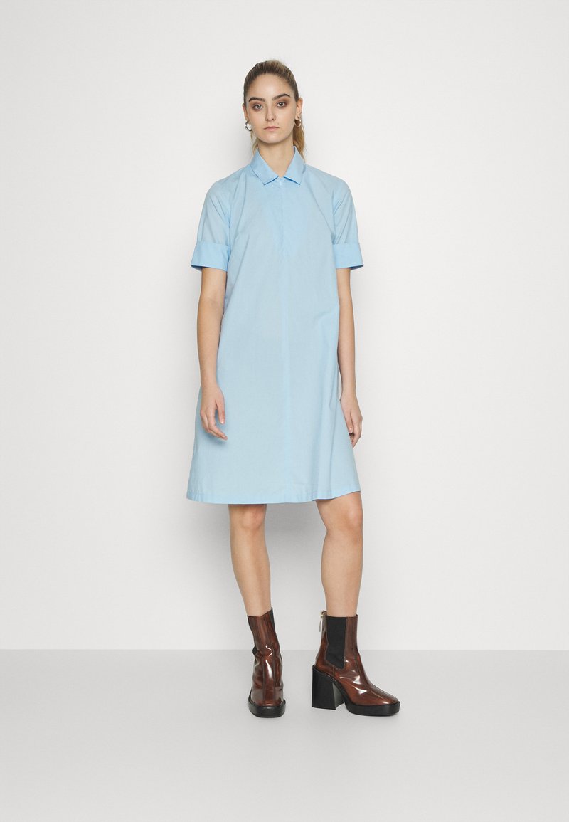 Vestido camiseiro azul claro com gola e mangas curtas, apresentando um corte reto e uma bainha até ao joelho. Usado com botas de salto brilhantes, castanhas.