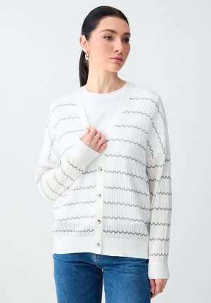 Donna con capelli scuri legati in una coda di cavallo, indossa un cardigan bianco a costine con strisce grigie ondulate e jeans blu, in piedi su uno sfondo semplice.