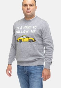 Maglione grigio lavorato a maglia con una grafica di un'auto gialla e il testo "È DIFFICILE SEGUIRMI", con polsini e orlo a costine. Indossato con jeans blu.