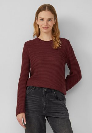 Vrouw met lichtbruin haar, gekleed in een gestructureerde bordeauxrode trui en zwarte spijkerbroek, staat tegen een grijze muur en glimlacht lichtjes.