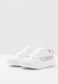 Zapatillas blancas de cuero sintético con punta redonda, suela texturizada, tacón con detalles dorados y el logo "TOMMY" exhibido de manera prominente.