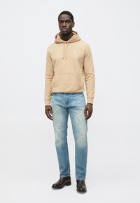Béžová mikina s kapucňou a predným vreckom, v kombinácii s vyblednutými modrými džínsami s fľakmi od farby a čiernymi členkovými topánkami.