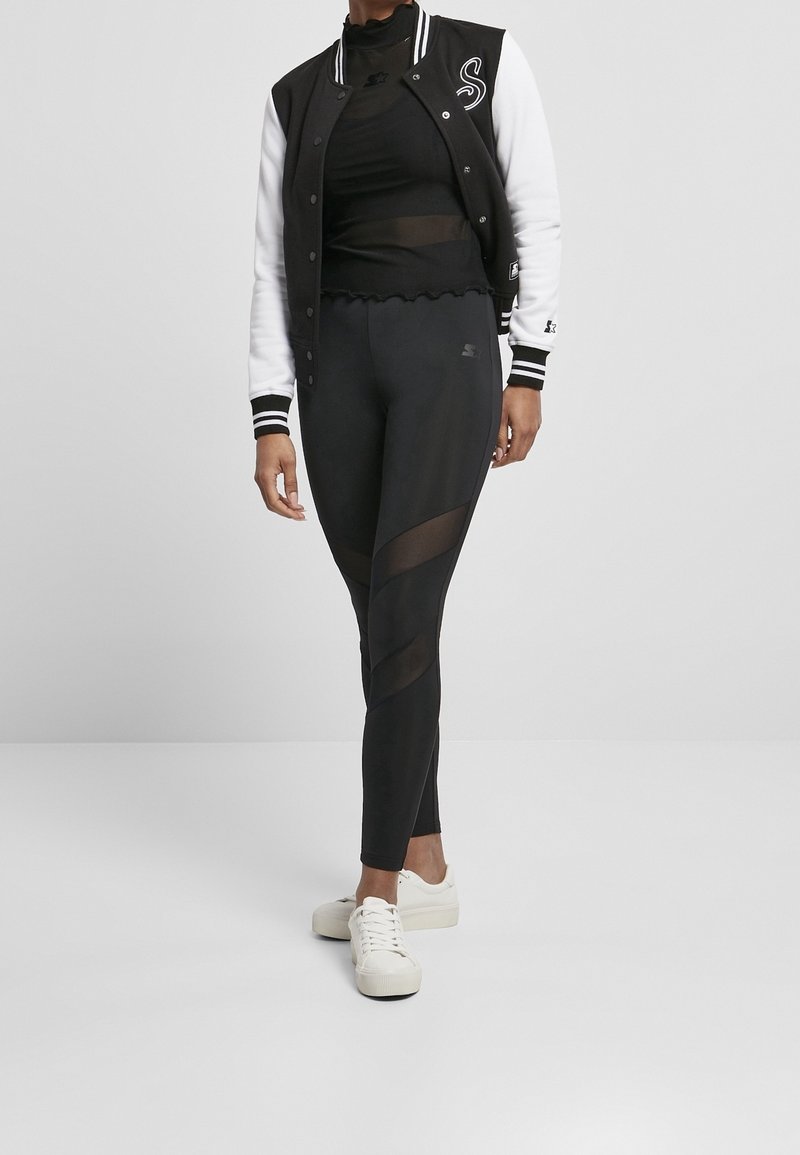 Femme portant une veste universitaire noire et blanche, un haut noir transparent, des leggings noirs avec des panneaux en maille, et des baskets blanches, se tenant sur un fond uni.