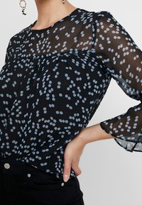 Schwarze transparente Bluse mit hellblauem Punktenmuster, runder Halsauschnitt und lockeren Ärmeln. Verfügt über geraffte Details und einen sanften Fall.
