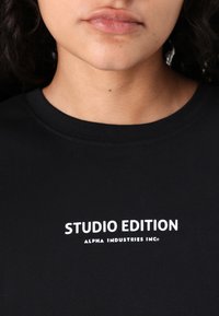Čierne tričko z mäkkej látky, s bielym potlačeným textom "STUDIO EDITION" a "ALPHA INDUSTRIES INC" na prednej strane, v priliehavom strihu.
