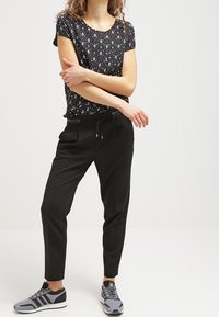 Mujer de pie con los brazos cruzados, vistiendo una camiseta negra con patrón, pantalones negros con cordón y zapatillas Adidas grises.