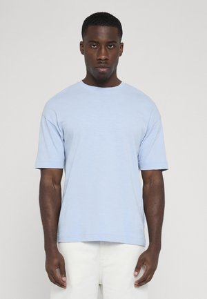 Hombre que lleva una camiseta de manga corta azul claro y pantalones blancos, de pie frente a un fondo liso de color gris claro.