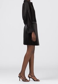 Blouse noire avec un col montant et des manches bouffantes, associée à une jupe noire plissée. Les chaussures sont à lanières avec un talon fin.