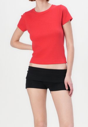 Kvinde iført en tætsiddende rød kortærmet top og sorte shorts med opfoldet linning, stående med den ene hånd bag ryggen.