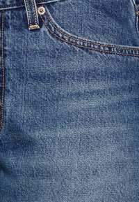 Gros plan sur un jean en denim bleu montrant la poche avant, la boucle de ceinture, le rivet en cuivre et les coutures orange visibles.