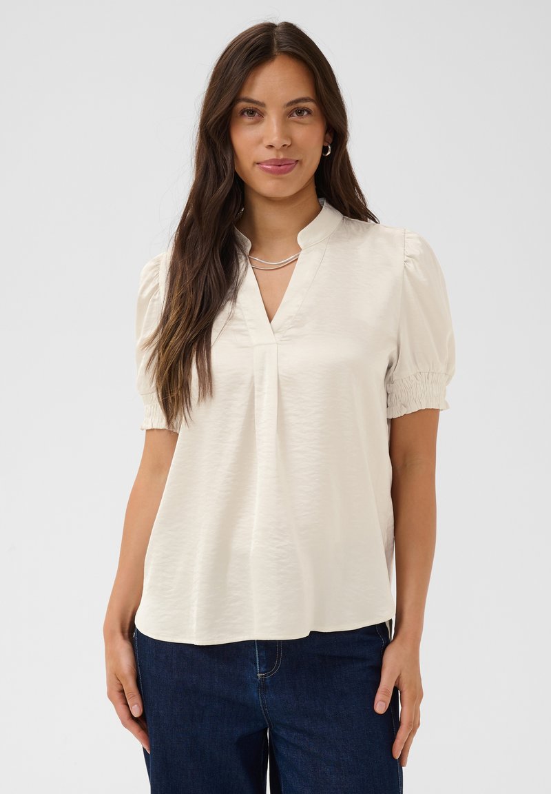 Femme aux longs cheveux foncés portant un chemisier crème avec des manches courtes bouffantes et un col en V, associé à un jean bleu foncé, fond neutre.