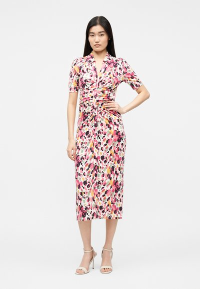 Femeie stând cu mâna pe șold, purtând o rochie de lungime medie cu mâneci scurte, cu pete abstracte roz, negre și galbene și sandale bej cu toc înalt.