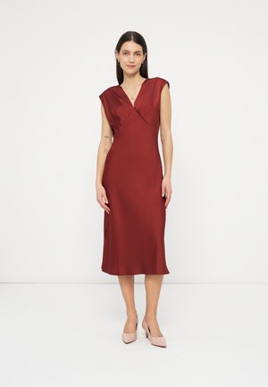 Žena stojí otočená smerom k fotoaparátu, oblečená v tmavomodrom midi šate bez rukávov a svetloružových lodičkách na jednoduchom bielom pozadí.