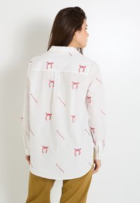 Chemise blanche avec un imprimé de ruban rouge, manches longues, design à boutons, ourlet arrondi et détail de pli au dos, portée avec un pantalon couleur beige.