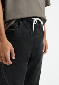 Czarne spodnie ze ściągaczem w talii, wyposażone w boczne kieszenie i wykonane z miękkiego, teksturowanego materiału. Ręka spoczywa na udzie. Prosty design.