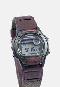 Relógio digital com caixa de plástico verde, bracelete de couro castanho, ecrã LCD a mostrar a hora e outras funções, incluindo botões para luz e alarme.