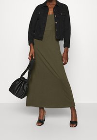 Olive zielona sukienka maxi o luźnym kroju, w połączeniu z czarną dżinsową kurtką i czarną skórzaną torebką-bucket. Czarnie klapki na obcasie dopełniają stylizację.