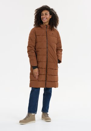 Bruine pufferjas met capuchon, horizontale stiksels, rits aan de voorkant en zijzakken. Gepaard met blauwe spijkerbroek en beige veterschoenen.