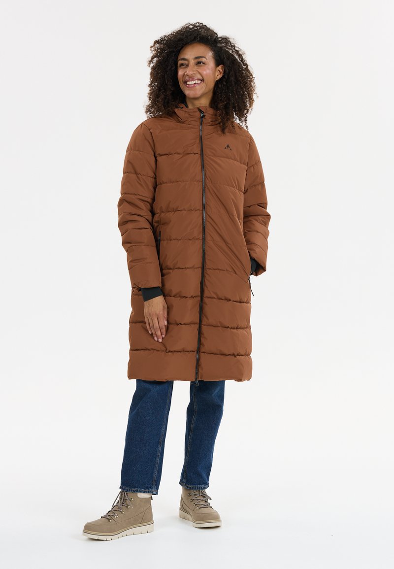Bruine pufferjas met capuchon, horizontale stiksels, rits aan de voorkant en zijzakken. Gepaard met blauwe spijkerbroek en beige veterschoenen.