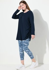 Tunic a maniche lunghe blu navy con arricciature laterali, abbinato a leggings blu con un motivo di onde. Il modello indossa sneakers bianche.