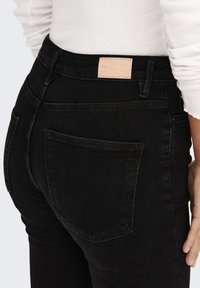 Jeans in denim neri a vita alta con una texture liscia, dotati di una tasca posteriore, cuciture a vista e un'etichetta in pelle rettangolare sul cinturino.