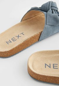 Blå ruskinds slidesandaler med en sløjfedetail, korkfodsvang og struktureret sål. "NEXT" logoet er synligt på indersålen.