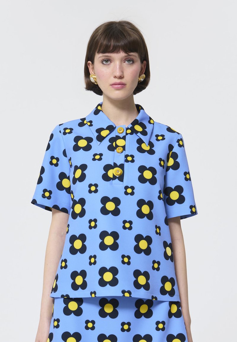 Blaue Blumenshirt mit kurzen Ärmeln, das große schwarze und gelbe Blumenmuster aufweist und drei gelbe Knöpfe an der vorderen Knopfleiste hat.