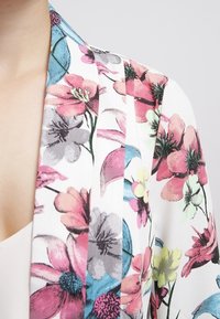 Lekka kurtka w kwiatowy wzór z intensywnymi różowymi, niebieskimi i szarymi kwiatami na białym tle. Projekt z kołnierzem szalowym.