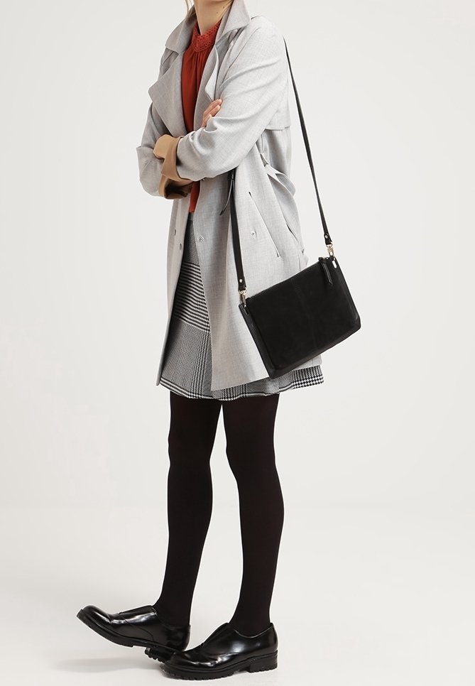 Osoba ubrana w szary płaszcz, wzorzystą spódnicę, czarne rajstopy, czarne mokasyny i nosząca czarną torbę na ramię na jednolitym tle.