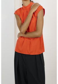 Top sans manches orange avec un col montant et des détails plissés, associé à un pantalon noir décontracté. Tissu lisse avec une texture légère.