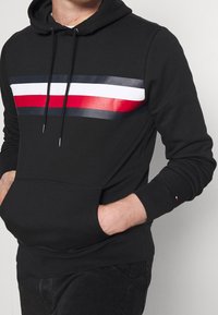 Sudadera negra con un patrón de rayas horizontales en rojo, blanco y azul marino en el pecho, con un bolsillo tipo canguro y capucha con cordón.