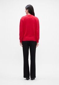 Rød strikket sweater med ribbestrikket kant og manchetter, parret med sorte skræddersyede bukser og højhælede sko, set fra ryggen mod en ensfarvet baggrund.