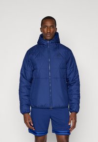 Mornarsko prešito jakno s kapuco, z sprednjim zadrgo in elastičnimi zapestji. Majhna črna ponarejena znamka na prsih. Oblečeno je z modrimi kratkimi hlačami.