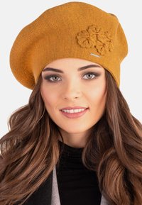 Senapsgul stickad beret med texturerad tyg, prydd med blommiga accenter och en liten metallisk logotyp på sidan.