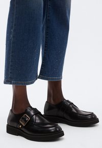 Fekete bőr cipők lekerekített orral és állítható csattal. Rövidített farmernadrággal viselve, amelyet kis szegecsek díszítenek.