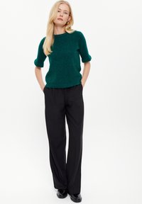Maglione verde a maniche corte con tessuto testurizzato, abbinato a pantaloni neri a gamba larga. Il modello sta in piedi con le mani nelle tasche, indossando scarpe nere.